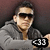 Pixelated image of a person wearing dark sunglasses and a gray jacket, holding a black object with white numerals.