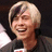 Pixelated close-up of a smiling person with short light-colored hair holding a microphone, wearing a dark jacket over a light shirt.