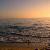 Pixelated sunset over water with gradient sky transitioning from blue to orange and yellow, featuring horizontal bands of color and blurred water reflections.
