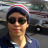 Pixelated image of a person wearing dark sunglasses, a dark blue jacket, and a cap with red and white stripes, standing in front of a blurred vehicle background.