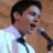 Pixelated close-up of a person with dark hair wearing a light-colored shirt against a brown background.