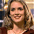 A pixelated close-up of a smiling person with light brown wavy hair, wearing a dark red top and a thin necklace, against a blurred background with warm tones.