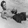 Black-and-white photograph of a shirtless man lying on a beach, holding a dark object near his chest.
