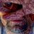 Pixelated static image with indistinct shapes in purple, brown, and blue tones, lacking clear subjects or text.