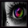 A close-up of a human eye with a rainbow-colored iris, black-and-white skin, and small black star and square tattoos on the cheek.