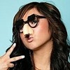 A static image of a person with long dark hair, wearing black-rimmed glasses, making a peace sign with their right hand against a light blue background.