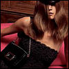 A woman with long brown hair wears a black strapless top and a black cap, holding a black handbag with a silver chain strap against a pink and brown background.