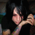Pixelated static image of a person with dark hair wearing black clothing, holding an object near their face against a dark background.