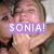 Two people embrace in a close-up; one wears green clothing, the other has light brown hair. A purple text box with 'SONIA!' appears over their faces.
