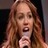 Pixelated close-up of a person with long brown hair holding a microphone, mouth open as if singing or speaking.