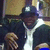 A person wearing a dark cap and jacket with white accents poses in front of a blurred background, captured across showing slight movement