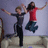 Two people jump on a beige couch against a light purple wall, with the right figure's arms raised higher in later frames.