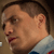 Pixelated close-up of a person's face in profile view, showing short dark hair, light skin tone, and a light-colored collared shirt with blue and red accents.