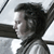Pixelated grayscale image of a person with dark hair in profile view, wearing a dark jacket over a light shirt, against a light background with dark diagonal lines.