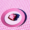 A pink and white checkered background with a circular pink-ringed plate holding a pink and brown striped object casting a dark blue shadow.