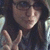 Pixelated image of a person with dark hair and light skin making a hand gesture, wearing dark clothing against a blurred background.
