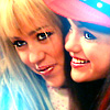 Two people with light brown hair and blue eyes pose closely together, one wearing a pink hat with blue and white accents.