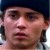 Pixelated close-up of a person's face with dark hair, wearing a dark blue cap and dark clothing, against a blurred background.