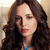 A pixelated close-up of a person with long dark brown hair, wearing a blue top and red lipstick, against a light brown background.