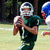 A football player in a green uniform and white helmet holds a ball, with another player partially visible behind him on a grassy field.