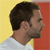 Pixelated profile view of a person with short dark hair, light skin, and a beard, wearing a white shirt against a yellow and red background.
