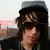A person with long dark hair and sunglasses wears a gray cap, standing against an outdoor background with buildings and sky.