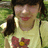 A person with dark hair and bangs wears a light yellow shirt featuring a red flower with a yellow center, standing outdoors on grass.