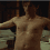A shirtless man with short brown hair stands in a dimly lit room with wooden bookshelves, shifting his pose