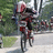 A person in red and white cycling gear rides a bicycle on a paved path with greenery in the background.