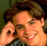 Pixelated static image of a person with brown hair and light skin smiling while holding hands near their face, wearing a dark top against a blurred green background.