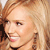 Pixelated close-up of a person with light brown hair, fair skin, and a slight smile, wearing a dark top with a visible collar.