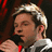 Pixelated close-up of a person with light brown hair, wearing a white shirt and red tie, holding a black microphone against a dark background with red accents.