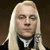 Pixelated close-up of a person with long straight blonde hair, wearing a black garment with a white collar, against a dark brown background.