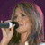 A person with light brown hair holds a dark microphone near their mouth against a purple background.
