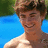 A pixelated image of a person with light brown hair smiling, wearing no shirt, against a blue water and green foliage background.