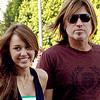 Two people stand outdoors with green foliage in the background; the person on the right wears dark sunglasses and a maroon shirt while the person on the left wears a dark blue sleeveless top with a green stripe.