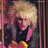 Pixelated image of a person with light-colored hair wearing dark clothing and pink accents, set against a blurred background.
