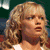 Pixelated close-up of a person with light brown curly hair, wearing a white top against a dark green background.