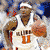 A basketball player in a white jersey with orange text and number 11 holds an orange ball, while another player in red is visible behind him.
