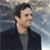 Pixelated static image of a person with dark hair wearing a dark jacket over a gray shirt against a blurred background with light and dark tones.