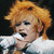 Pixelated close-up of a person with voluminous orange hair holding a white microphone against dark background.
