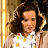 Pixelated static image of a person with curly dark hair wearing a white shirt with yellow and red accents, set against a warm-toned background.