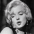 Black-and-white pixelated image of a person with light-colored hair styled in waves, mouth open in mid-expression, wearing a dark garment with a light collar.