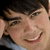 Pixelated close-up of a person with dark hair, light skin, and a wide smile showing teeth, wearing a light blue garment against a dark background.