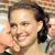 A pixelated close-up of a person with dark hair tied back, smiling broadly with visible teeth, holding a microphone near their mouth against a blurred background.