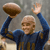 A person in a blue long-sleeve shirt holds a brown football above their head with one hand while raising the other hand in a wave gesture against a gray background.