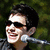 A person with short dark hair and sunglasses smiles while holding a silver microphone against a blurred green background.
