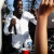 Pixelated figure in white shirt and dark pants holding a microphone near mouth, standing beside tall brown structure with blurred background.