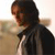 A pixelated figure with dark hair in a ponytail, wearing a dark jacket over a light shirt, viewed from the side against a blurred background.