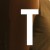 A white capital letter T centered on a blurred brown and orange gradient background with dark vertical shadows.